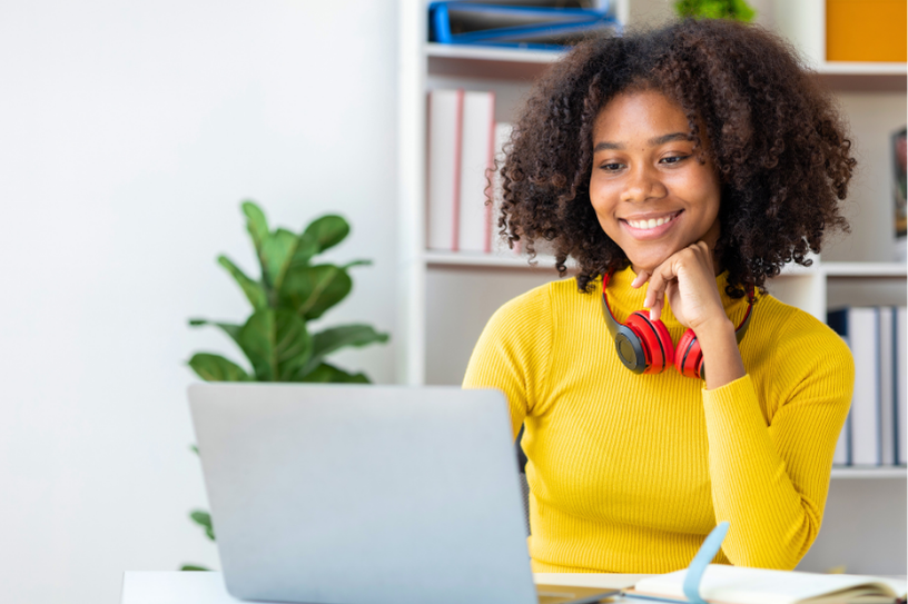 A teen girl wearing a yellow sweater and red headphones smiles while looking at a laptop screen.