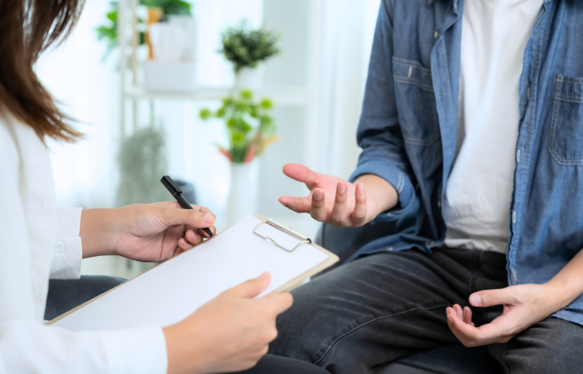 A person in a white shirt takes notes on a clipboard while conversing with another person in a denim shirt.