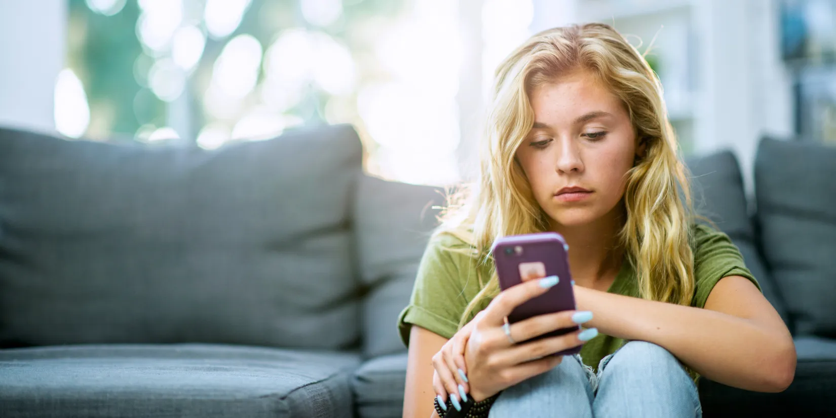 A teen girl sitting on the floor looking at a smartphone with a serious expression.