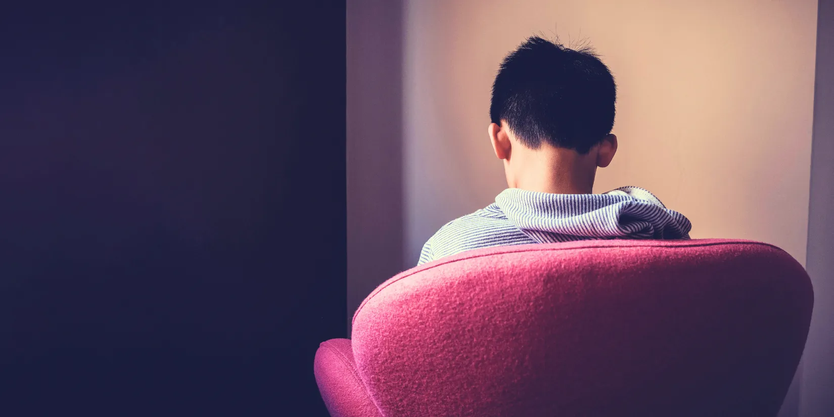 Boy sitting in a pink chair looking down.