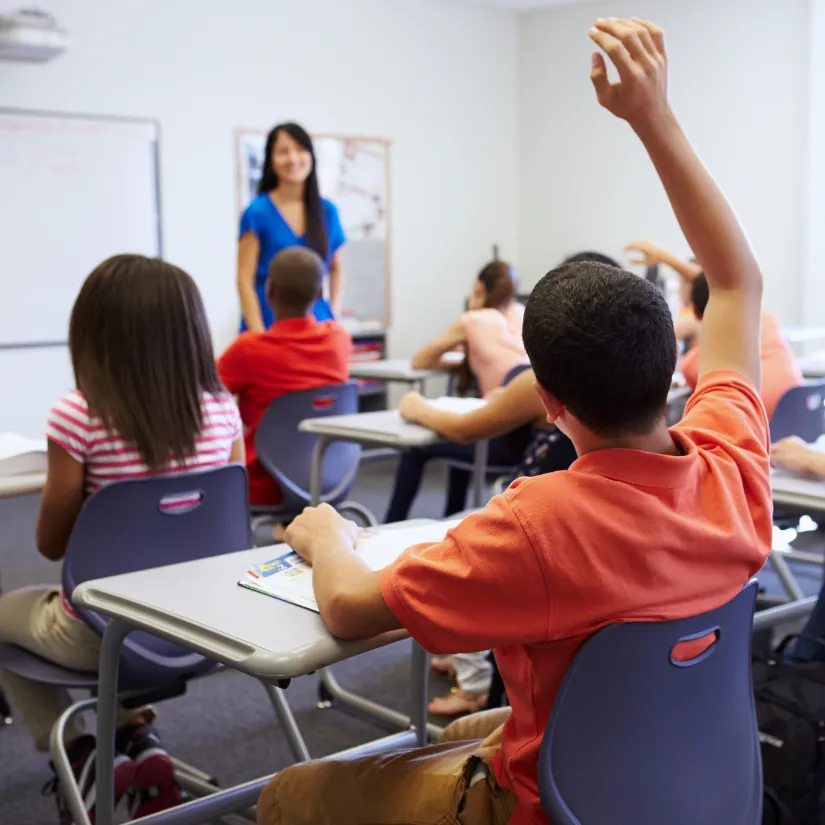 A teacher stands at the front of a classroom as students sit at their desks, with one student raising their hand.