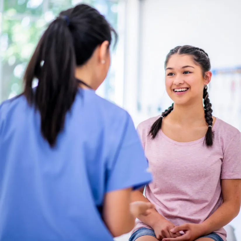 A healthcare professional in a blue scrub top talks to a young person with braided hair sitting in a clinic examination room.