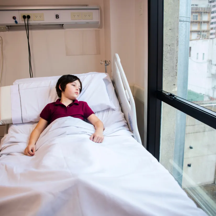 A young person lies in a hospital bed looking out a window at buildings.