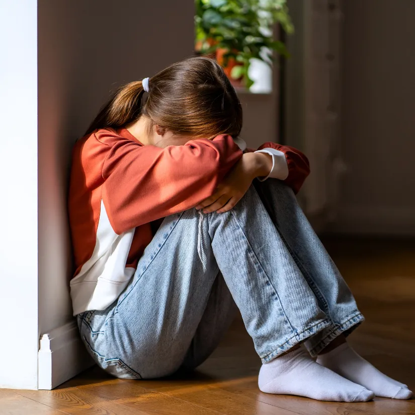 A youth sits curled up on the floor with their head in their arms, illuminated by sunlight streaming in from a window.