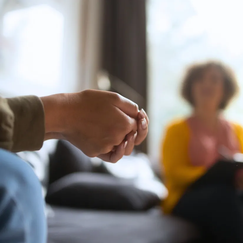 Close-up of clasped hands during a counseling session, with a blurred figure in the background holding a tablet.