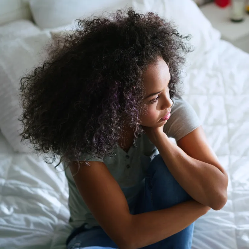 A teen with curly dark hair sits on a white bed, hugging their knees and looking away from the camera.